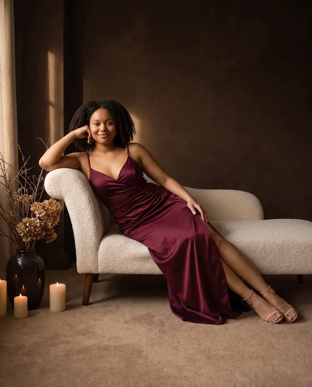 Maya in the wine satin Celeste dress, reclining on a cream chaise